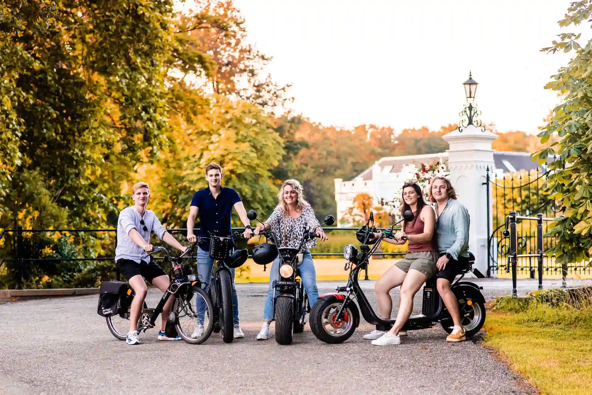 Vijf vrienden met een Solex en e-Choppers bij een wit toegangshek in de herfst